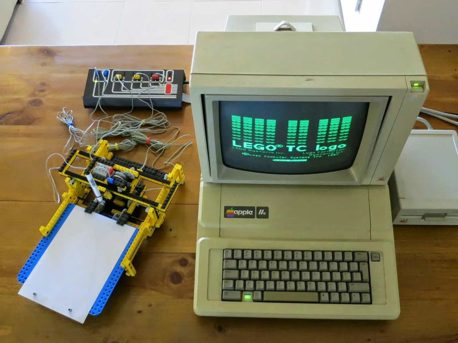 Un plotter de LEGO con un rotulador sobre una hoja, conectado a la interfaz de LEGO TC Logo, junto a un Apple IIe.